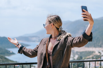Travel blogger filming live stream on smartphone against mountain coastline, Gen Z female tourist on viewpoint, panoramic sea and hills, social media content creator, sunny outdoor vacation vibe. © shintartanya