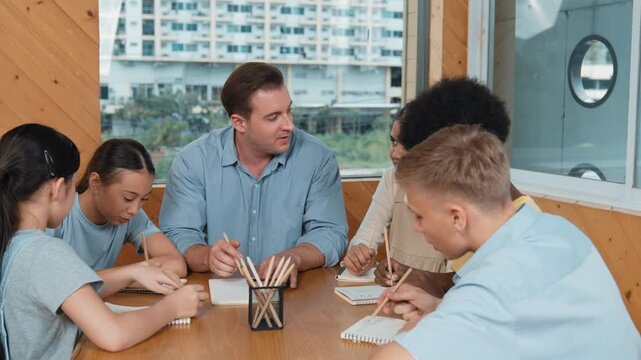 Smart teacher talking and explaining work while multicultural children drawing or taking a note in paper at table with pencil. Happy diverse student drawing or sharing idea while sitting. Edification