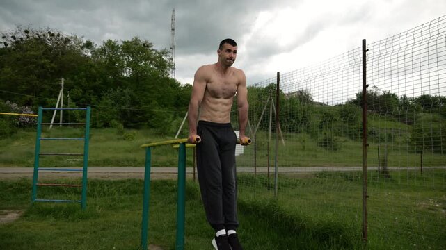 Strong man trains abdominal muscles raising legs on parallel bars at sports ground. Young shirtless sportsman exercising at nature. Sporty athlete working out at nature. Concept of sportive lifestyle