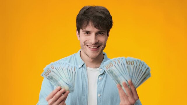 A young man stands in a studio holding multiple bundles of cash while facing the camera. The background is bright orange, emphasizing his focused expression and the money he displays.