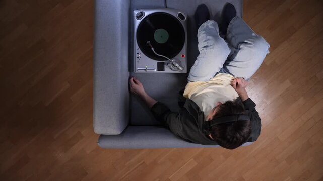 Young adult lying on couch, relaxing and listening to vinyl on a turntable with headphones, enjoying a peaceful, cozy moment of analog music and weekend escape at home