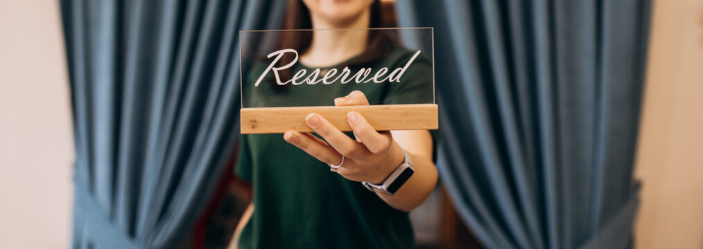 Unrecognizable woman holding Reserved sign on outstretched hand. Focus on table reservation plaque with blue velvet curtains backdrop. Hospitality concept.