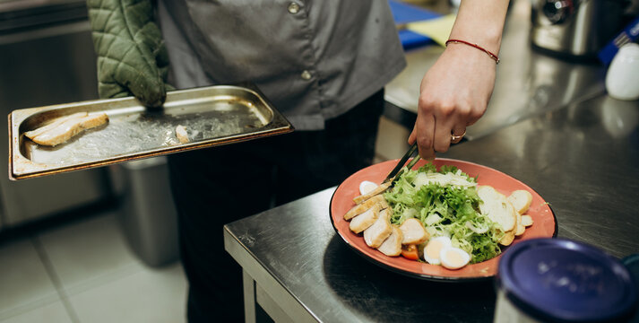 Close up of chef placing cooked chicken fillet onto plate while preparing salad. Professional restaurant kitchen food plating process.