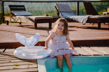 A little girl is sitting on the pool in a mermaid costume. Beautiful happy girl of preschool age resting. Inflatable swimming ring with fish tail. Warm and sunny weather.