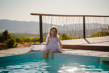 A preschool girl sits on the edge of the pool and wets her feet in the water. Happy girl relaxing in the pool with a swimming circle. Good warm weather, setting sun.