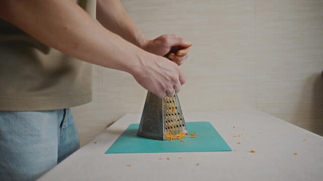 Medium close-up of a man grating carrot with one hand on a kitchen grater. Simple food preparation and everyday home cooking routine without showing face. High quality 4k footage