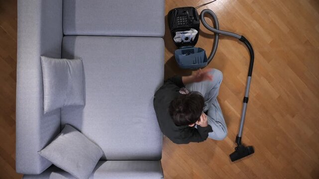 Man sitting on wooden floor beside gray sofa, dismantling a vacuum cleaner while talking on his mobile phone during a break from household chores in a bright apartment