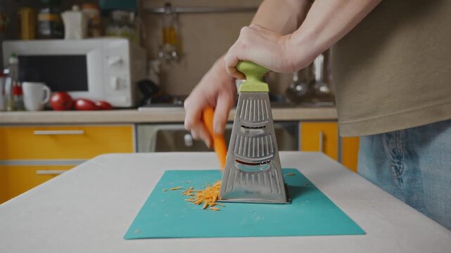 Medium shot of a man grating fresh carrot on a kitchen grater at home. Everyday food preparation process, healthy cooking and simple domestic lifestyle without showing face. High quality 4k footage