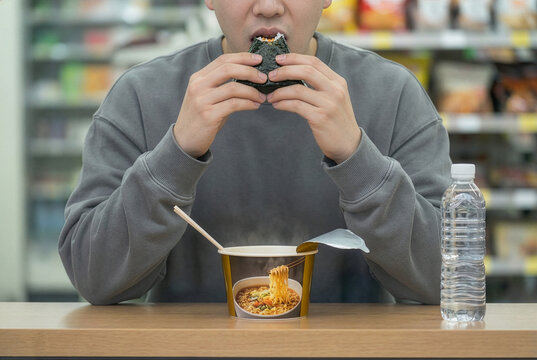 young man eating triangular Kimbap with ramyeon in a convenient store