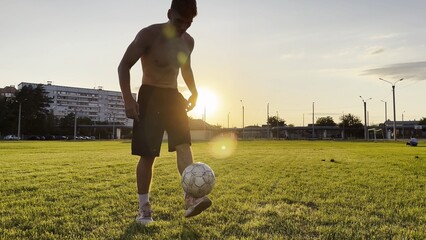 Young man juggling soccer ball on stadium at sunset. Professional footballer kicking ball at green field. Sportsman practicing tricks at meadow with sunlight at background. Freestyle football