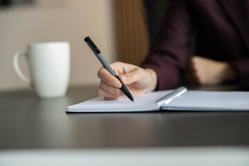 Close-up of a woman's hand holding a pen and writing something down on a notepad in a home office. A cup of coffee is next to it. © Dejan