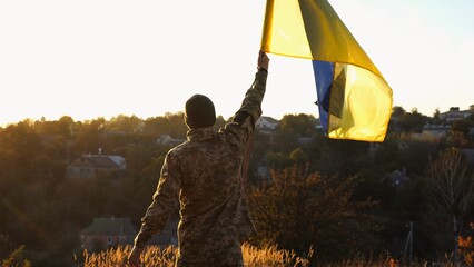 Young soldier of ukrainian army lifted blue-yellow banner in honor of the victory against russian aggression at sunset. Male military in uniform raised a waving flag of Ukraine at countryside