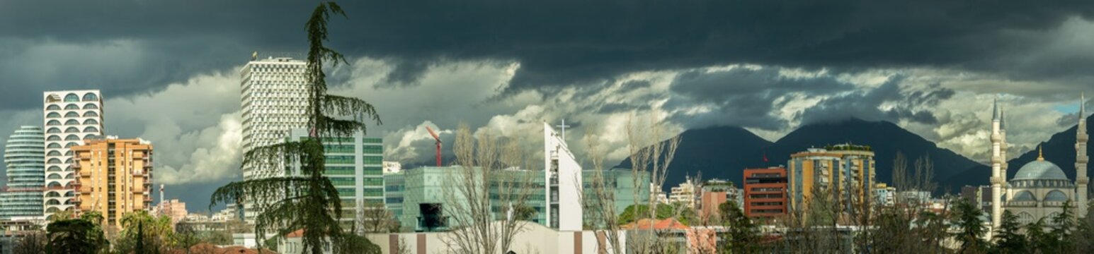 A wide-angle panoramic view of the Tirana city skyline under a heavy, dramatic cloud cover. The shot captures the architectural evolution of the Albanian capital, featuring the Scanderbeg Head tower (