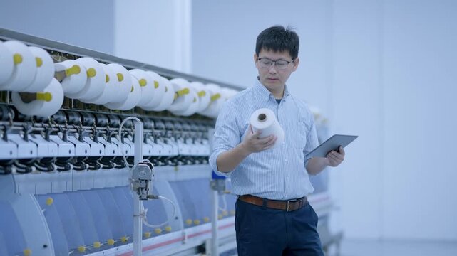 Young male engineer in a factory workshop holding a yarn spool while inspecting machinery