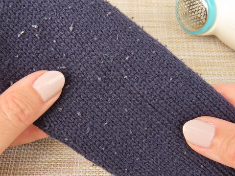 Closeup of woman hands holding dark blue sweater with pills. Sweater cuff with pills, also known as lint balls or fuzz. Electric fabric shaver and lint remover on the background.