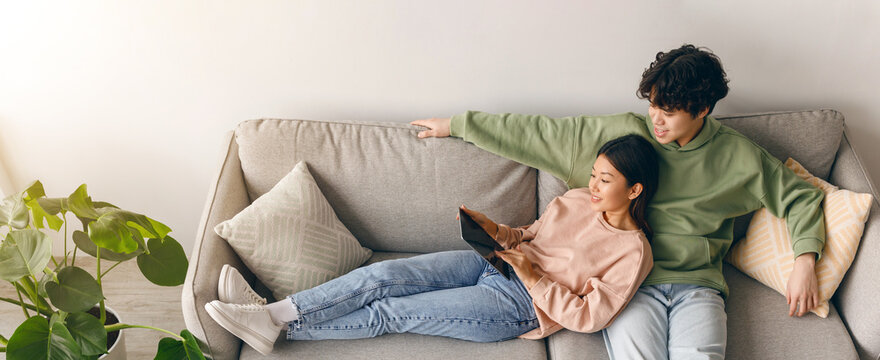 A young couple sits on a sofa in a living room. They share a tablet, looking at the screen. One person holds the tablet while the other leans back comfortably. Green plants add to the scenery.