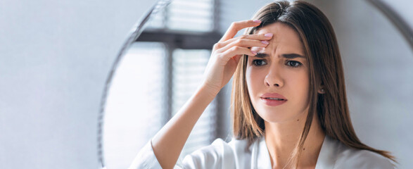 A woman stands indoors in a well-lit room with a worried expression. She holds her forehead with one hand as if feeling stress or discomfort. The atmosphere suggests a common moment of concern. © Prostock-studio