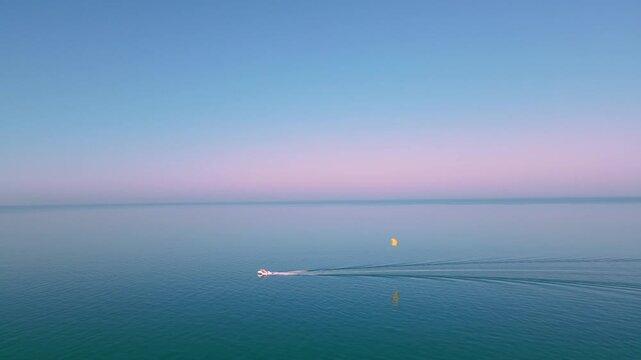 Small boat towing yellow parasail across calm blue sea at sunset. Relaxed holiday mood, 4K cinematic coastal footage.