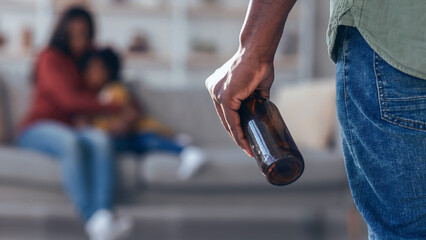 A man stands with a bottle in his hand. In the background, a woman and a child are seated on a sofa. They are engaged in a quiet moment together in their home. © Prostock-studio