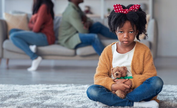 A young girl sits on the floor holding a teddy bear. Her parents are seated on a couch, facing away from each other, showing conflict. The scene captures a moment of tension in a home.