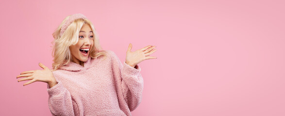 Fototapeta premium A girl with blonde hair is smiling widely while wearing a pink sweater. She has her hands raised in excitement. The background is a soft pink color.