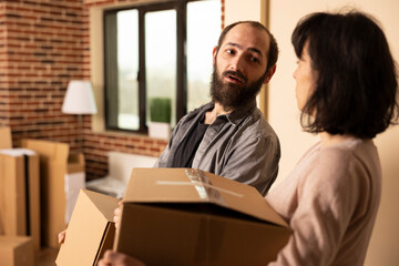 Fototapeta premium Caucasian husband and wife carrying belongings into brick wall apartment. White couple entering living room with moving boxes, chatting while relocating into newly purchased home.