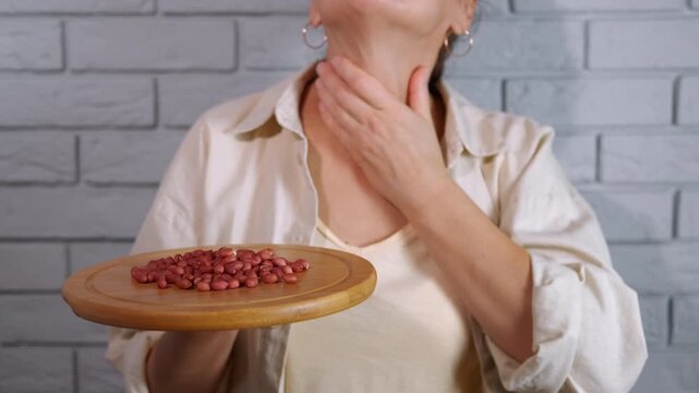 Woman with a peanut allergy touching her throat. Female individual suffering from an allergic reaction, holding a wooden plate with peanuts while touching her throat in distress due to swelling