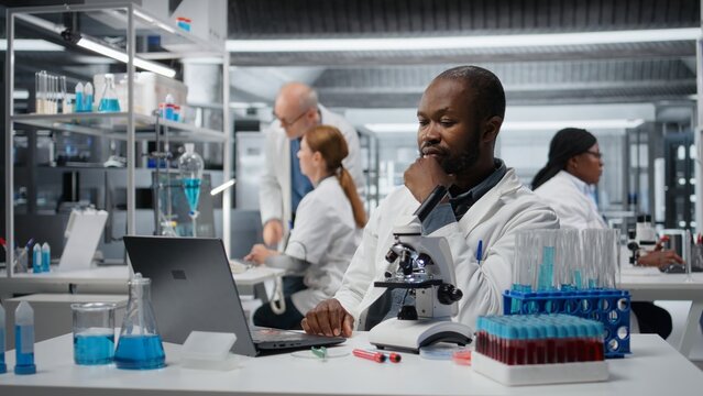 Stressed lab chemist feeling overwhelmed while working on laptop, studying genomics. Anxious African american man struggling with results while studying DNA strand mapping, camera A
