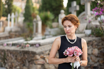 Fototapeta premium Sorrowful woman in mourning black dress standing in cemetery with flowers in hands against backdrop of tombstones and stone crosses on summer day