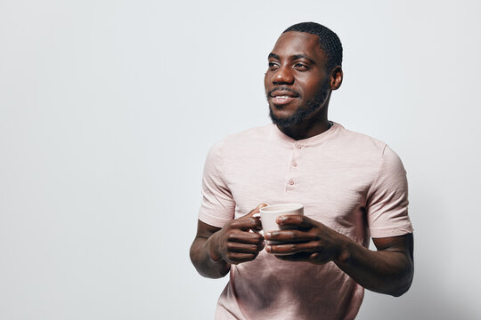 African American man with dark skin holding white cup, smiling and looking away. Casual clothing, short hair, standing against plain light background with relaxed and happy expression.