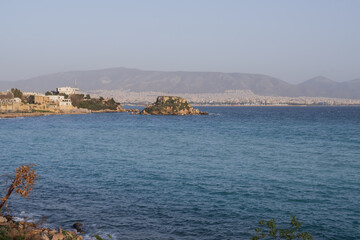 Wide view of Athens coastline showing blue sea, rocky islet and city buildings in the distance. Peaceful Mediterranean seascape for travel themes. © Olha