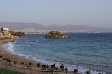 Wide view of Athens coastline showing blue sea, rocky islet and city buildings in the distance. Peaceful Mediterranean seascape for travel themes. © Olha