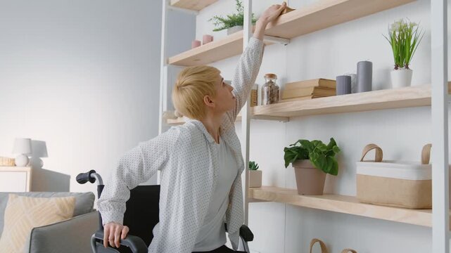 Problems of life with disability. Calm mature woman wheelchair user reaching up near shelves and taking photo album, watching photographies at home, zoom in portrait, slow motion