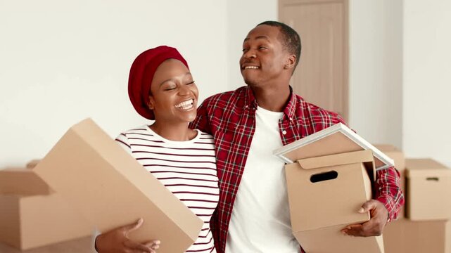 An African American woman wearing a red head wrap and a striped top stands next to an African American man in a plaid shirt. They hold boxes and look happy while moving into their new home.