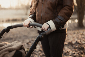 Fototapeta premium A woman in a brown aviator jacket holds her hands on the handle of a baby carriage while walking in the park at sunset