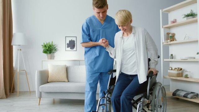 Successful rehabilitation concept. Professional man therapist helping mature woman to stand up from wheelchair and walk, getting healthy after accident at home, slow motion, free space