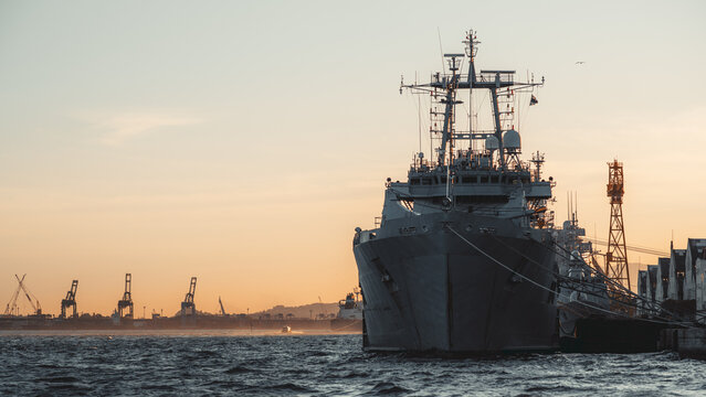 Front view of gray naval ship moored at industrial harbor at sunset, choppy sea foreground, port cranes and skyline in distance, warm golden sky with copy space