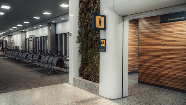 Modern airport terminal interior with empty waiting area seating, women restroom entrance and accessibility signage on pillar, wood panel wall, vertical garden, clean public space, travel concept