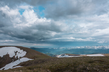 White snowfield on stone hill and large snowbound mountains on horizon under gray cloudy sky. Dramatic view from stony top to forest hills and big snowy mountain range far away in gloomy rainy weather © Daniil