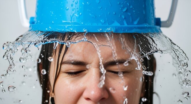 A person flinches as a blue bucket filled with water pours down onto their head in a studio setting.