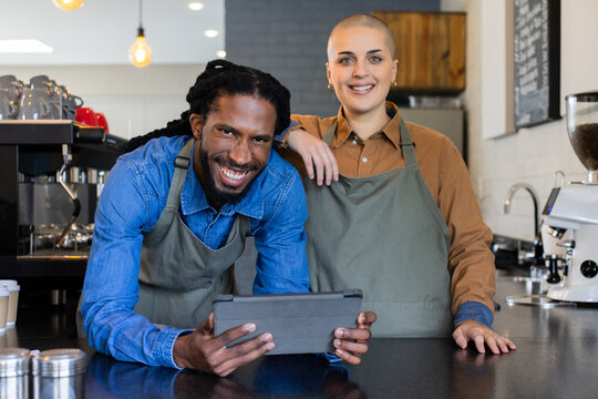 Diverse baristas leaning on counter holding tablet in coffee shop with espresso machine, paper cups