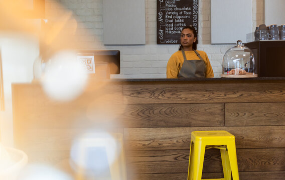 Indian woman barista standing behind cafe counter with pastry dome preparing coffee orders