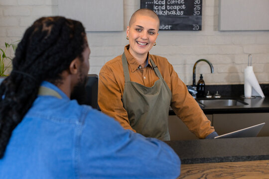 White woman barista smiling at African American man customer, using POS at counter