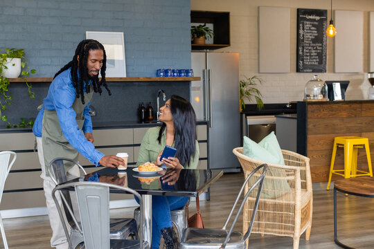 African American server in green apron handing coffee cup to Asian woman holding smartphone at cafe