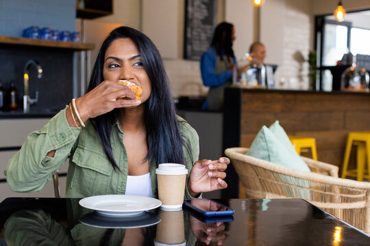 Asian Indian woman biting pastry and holding coffee cup at glossy black cafe table with smartphone
