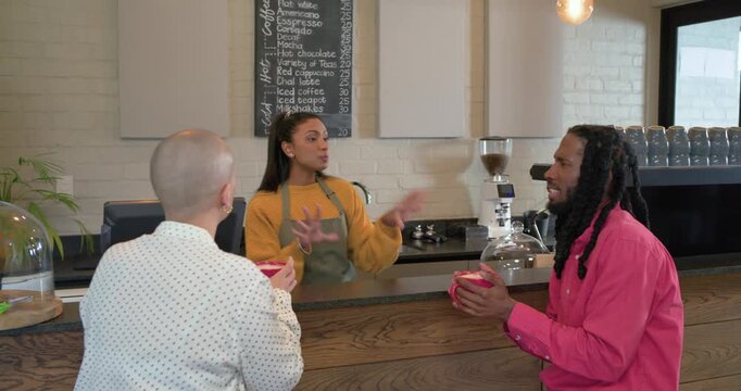 Indian barista greeting couple sipping mugs prompts explaining chalkboard menu in cafe and laughing