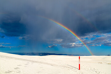 Naklejka premium Alkali Flat Trail in White Sands National Park, New Mexico. Scenic view features a vivid rainbow over white gypsum dunes and a red trail marker under a stormy sky
