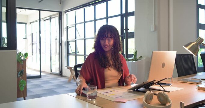 African American woman typing on laptop in office, grabbing glass, picking up pink phone, smiling
