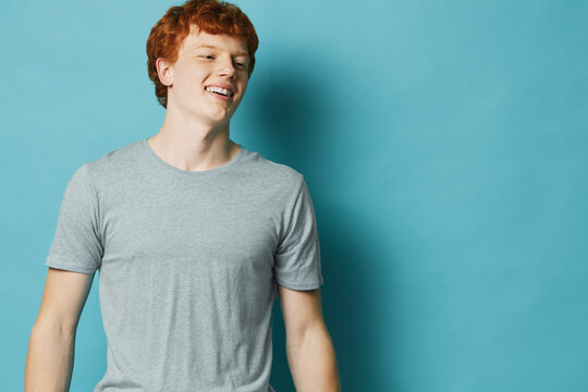 Young man with red hair smiling and wearing gray t shirt against blue background in studio. Casual portrait of happy male model with light skin and short hairstyle.
