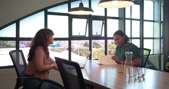 Women colleagues reviewing papers at conference table, left leaning, gesturing; right turning pages